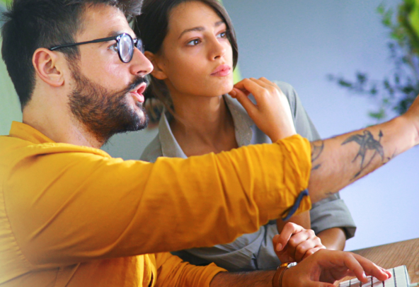 A man wearing a yellow shirt and glasses is sitting at a desk with a woman. The man is pointing at a screen and explaining something.