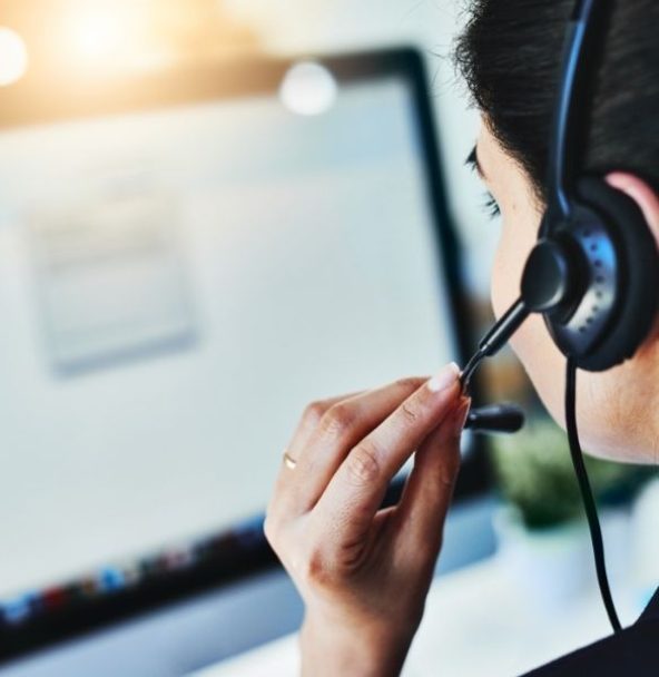 Person with headset and microphone, presumably in a conversation or call, sitting in front of a slightly blurred computer screen and adjusting the microphone with their hand.