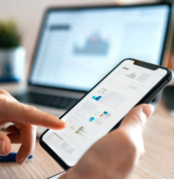 The image shows a smartphone. A hand is operating the smartphone. A laptop can be seen in the background.