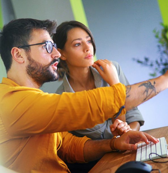 A man in a yellow sweater shows a woman something on his screen. The two are talking.