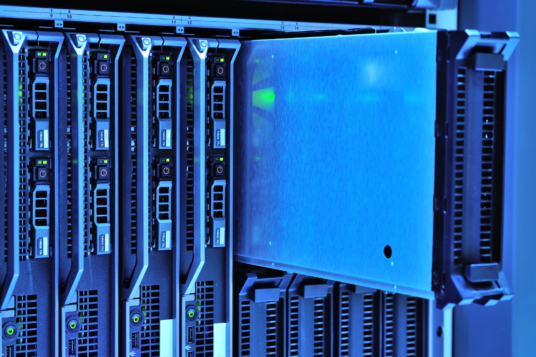 A close-up of a server rack glowing with blue light, with one server blade partially pulled out, revealing rows of blinking status lights and USB ports on the adjacent server blades.