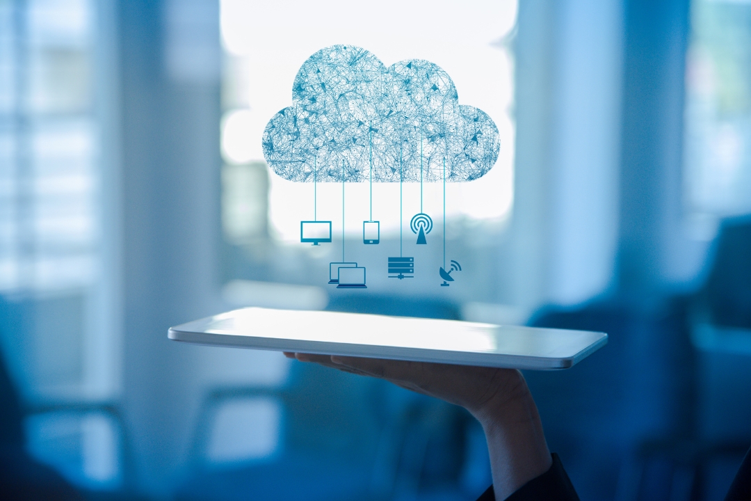 A person holds a white tablet horizontally, and above it floats a light blue, networked cloud icon. Various digital device icons—including a monitor (desktop), a smartphone, a tablet, and a laptop—are shown being serviced or provisioned directly from the cloud, symbolizing Virtual Desktop Infrastructure (VDI) delivery.