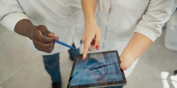 An overhead view of two people in white lab coats, representing developers or testers, collaborating while looking at a digital tablet. One person is pointing at the screen, symbolizing the review and analysis of code, data, or application functionality within a shared Development and Test Environment.