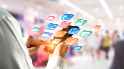 
A person is holding a smartphone with a bright orange glow, and several digital application icons (like a shopping cart, a cloud, a globe, and a video player) are floating out and around the screen against a blurred background of a public space.