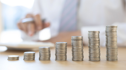 
A series of seven stacks of silver coins, arranged to look like a bar chart with an upward trend, sit on a wooden surface. In the background, a person in a white shirt and tie is blurred while working with a calculator.