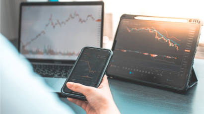
A person views financial or stock market charts with downward trends on three different screens: a laptop in the background, a tablet on a stand, and a smartphone held in the foreground. Sunlight streams in from the right.