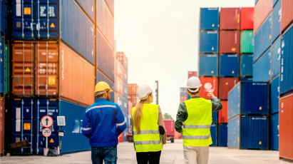 Three individuals wearing safety vests and hard hats (two yellow and one white) walk through a shipping yard, surrounded by tall stacks of multicolored cargo containers.