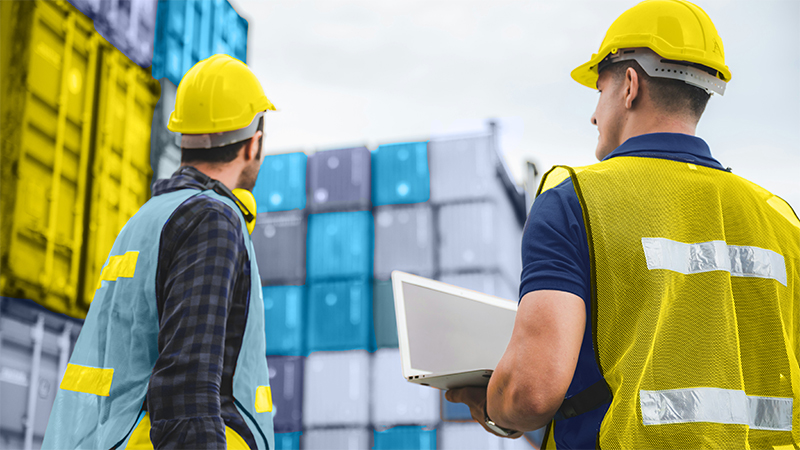 Three individuals wearing safety vests and hard hats (two yellow and one white) walk through a shipping yard, surrounded by tall stacks of multicolored cargo containers.