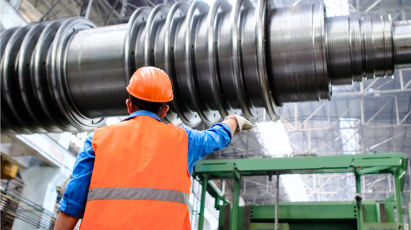 A manufacturing or industrial worker, wearing an orange safety vest and hard hat, stands with their back to the viewer, pointing up towards a massive, cylindrical piece of metallic machinery or turbine rotor in a large factory setting.