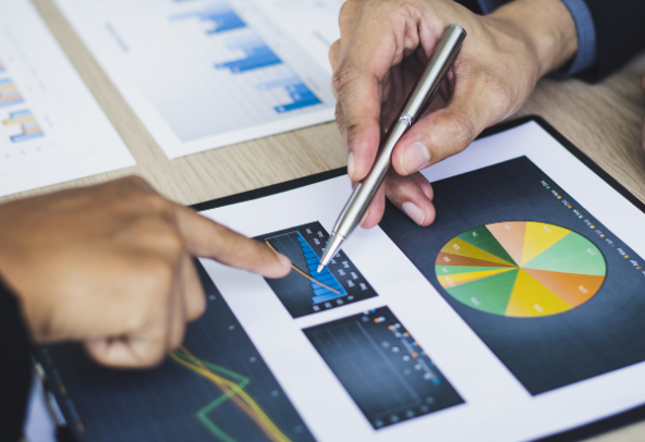 Business Continuity Management Microsoft The image shows two people reviewing printed business charts and graphs on a desk. One person is pointing at a bar chart, while the other is holding a pen and pointing at the same area. The documents include various types of charts, such as bar graphs, line graphs, and a colorful pie chart, suggesting a discussion or analysis of financial or performance data.