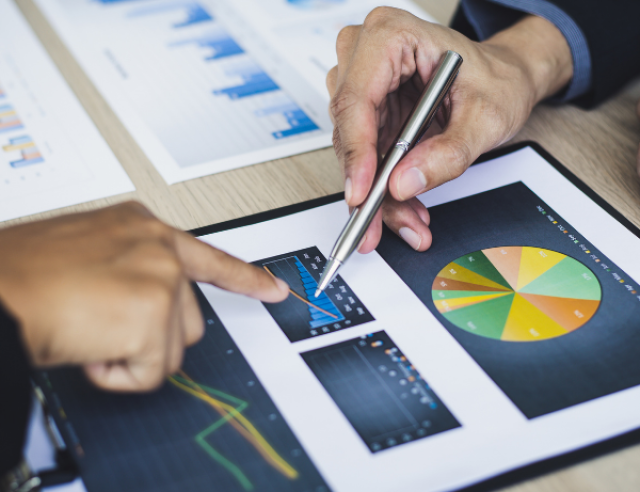 The image shows two people reviewing printed business charts and graphs on a desk. One person is pointing at a bar chart, while the other is holding a pen and pointing at the same area. The documents include various types of charts, such as bar graphs, line graphs, and a colorful pie chart, suggesting a discussion or analysis of financial or performance data.