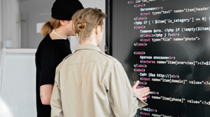 Two software developers stand looking at a large monitor displaying application code, with one person pointing and discussing the content. This represents a software development scenario where an AI-powered Google AgentSpace could serve as an expert assistant for coding, troubleshooting, or understanding legacy systems.