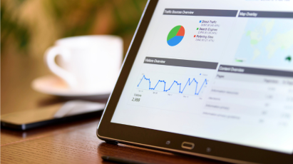 Dashboards & KPIs A close-up of a tablet screen displaying a digital analytics dashboard with a traffic sources pie chart, a website visitors line graph, and a map overview. A white coffee cup is blurred in the background, suggesting business analysis or reporting.