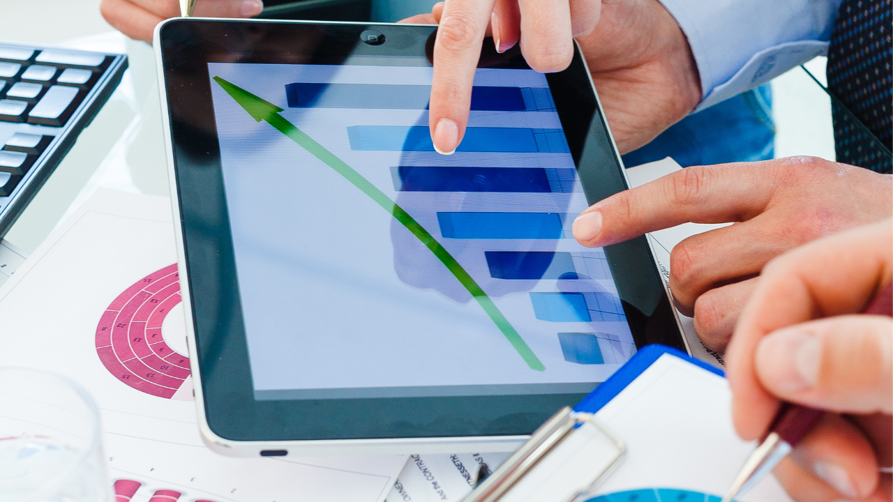 Tablet with a bar chart and a green downward-pointing arrow; several hands are pointing at the screen, with documents and another device in the background.