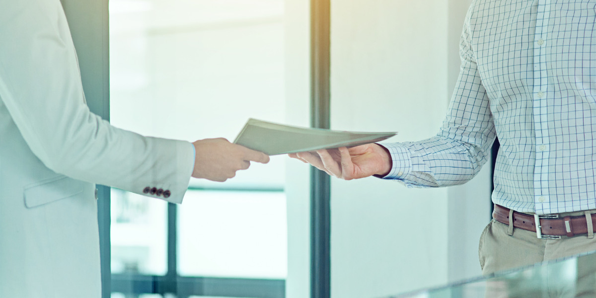 Zwei Personen tauschen ein Dokument aus; eine trägt einen hellen Anzug, die andere ein kariertes Hemd und eine beige Hose. Im Hintergrund ist ein modernes Büro mit Glaswänden und Tageslicht zu sehen.