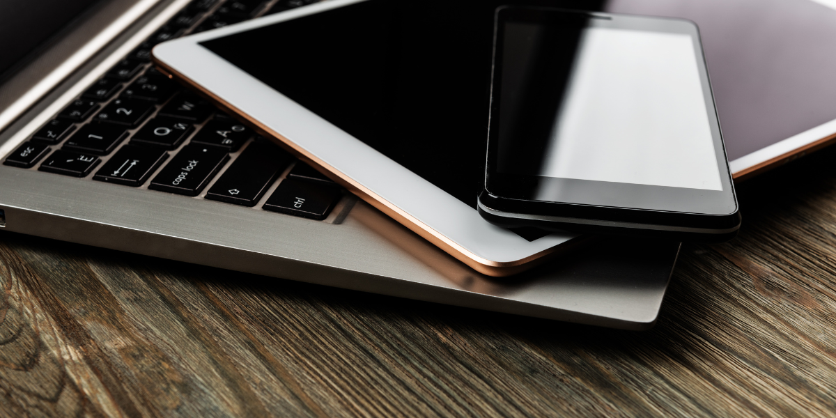 Close-up of a laptop keyboard with a tablet and a smartphone placed diagonally on top of the laptop; all devices are on a wooden surface.