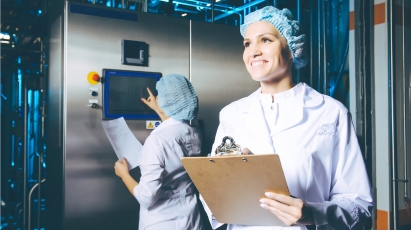 Product Analytics Two female food or pharmaceutical production workers in white lab coats and hairnets are in a clean manufacturing facility. The person in the foreground smiles while holding a clipboard, and the person behind her interacts with a digital control panel on a piece of large machinery.