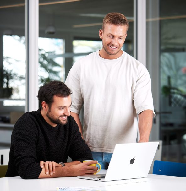 Zwei Männer arbeiten gemeinsam an einem Tisch; einer sitzt am Laptop und tippt, während der andere daneben steht und auf den Bildschirm schaut.