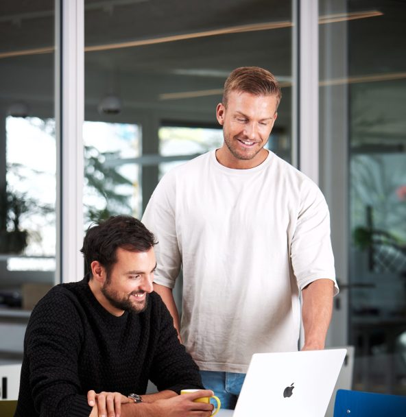 PCG - Security und Compliance 24 Zwei Männer arbeiten gemeinsam an einem Tisch; einer sitzt am Laptop und tippt, während der andere daneben steht und auf den Bildschirm schaut.