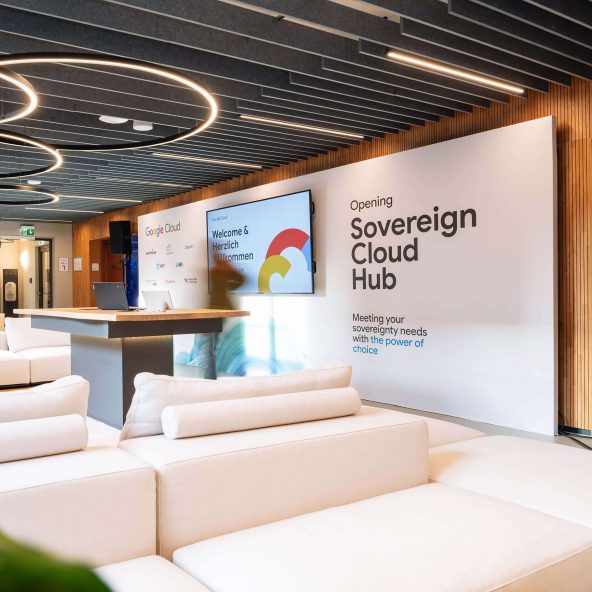 An image of a modern office space, likely a reception or event area, with large, comfortable white modular couches. A central white wall features the text "Opening Sovereign Cloud Hub Meeting your sovereignty needs with the power of choice." To the left, a monitor displays the Google Cloud logo and the welcome message "Welcome & Herzlich Willkommen." The ceiling has dark acoustic panels and illuminated circular light fixtures.