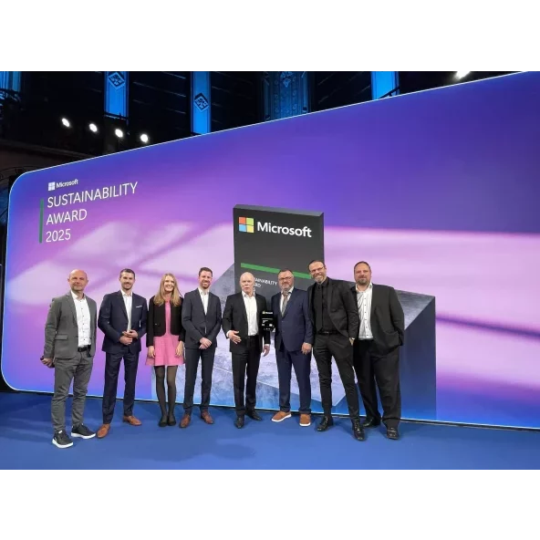Group of seven people, a mix of men and women in business attire, posing on a stage in front of a large screen displaying the Microsoft Sustainability Award 2025 logo and a large 3D trophy prop.