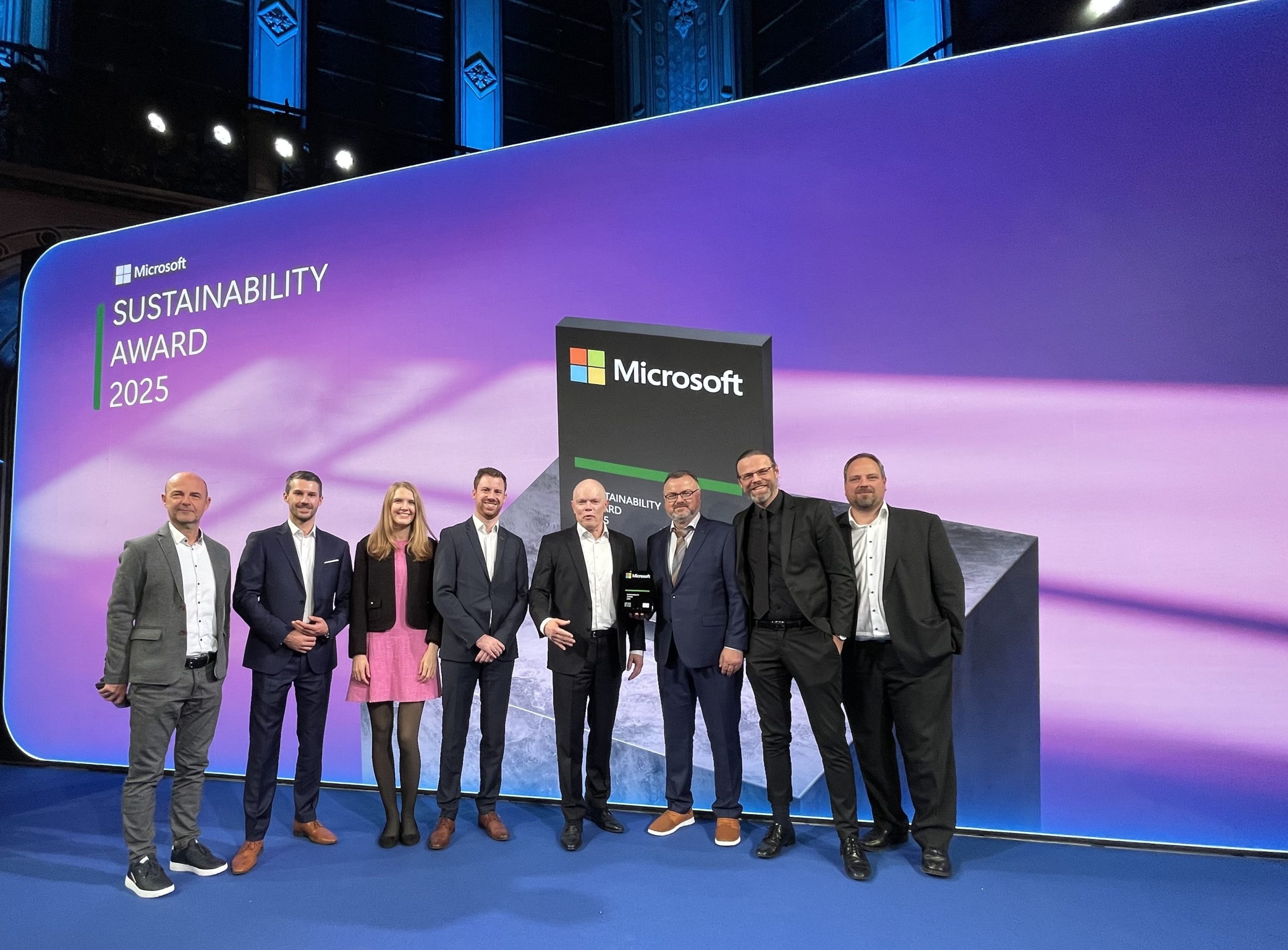 Group of seven people, a mix of men and women in business attire, posing on a stage in front of a large screen displaying the Microsoft Sustainability Award 2025 logo and a large 3D trophy prop.